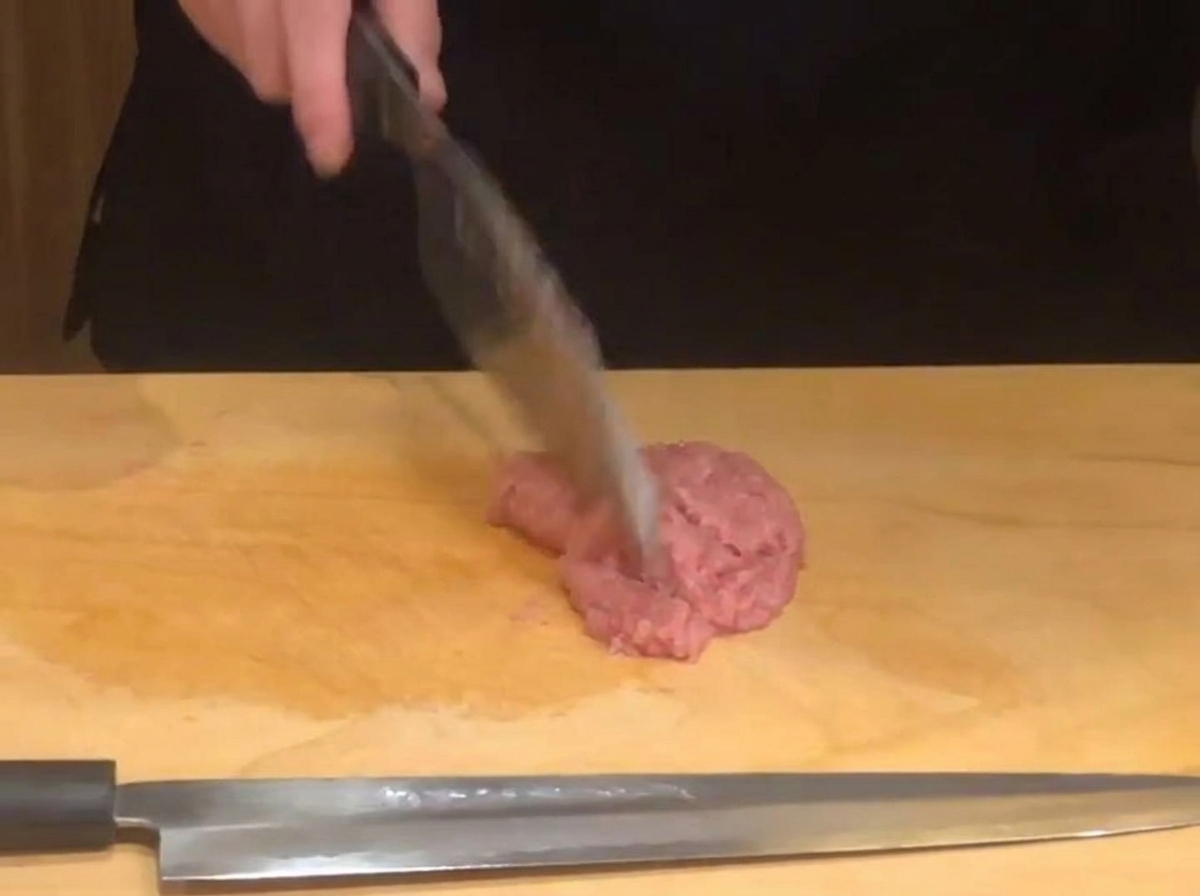 A chefs hand using a sharp knife to mince pink raw tuna on a wooden cutting board.