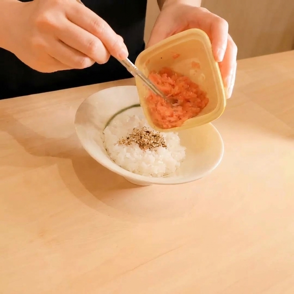 Minced raw tuna being scooped from a small plastic container onto a bed of white rice sprinkled with sesame seeds.