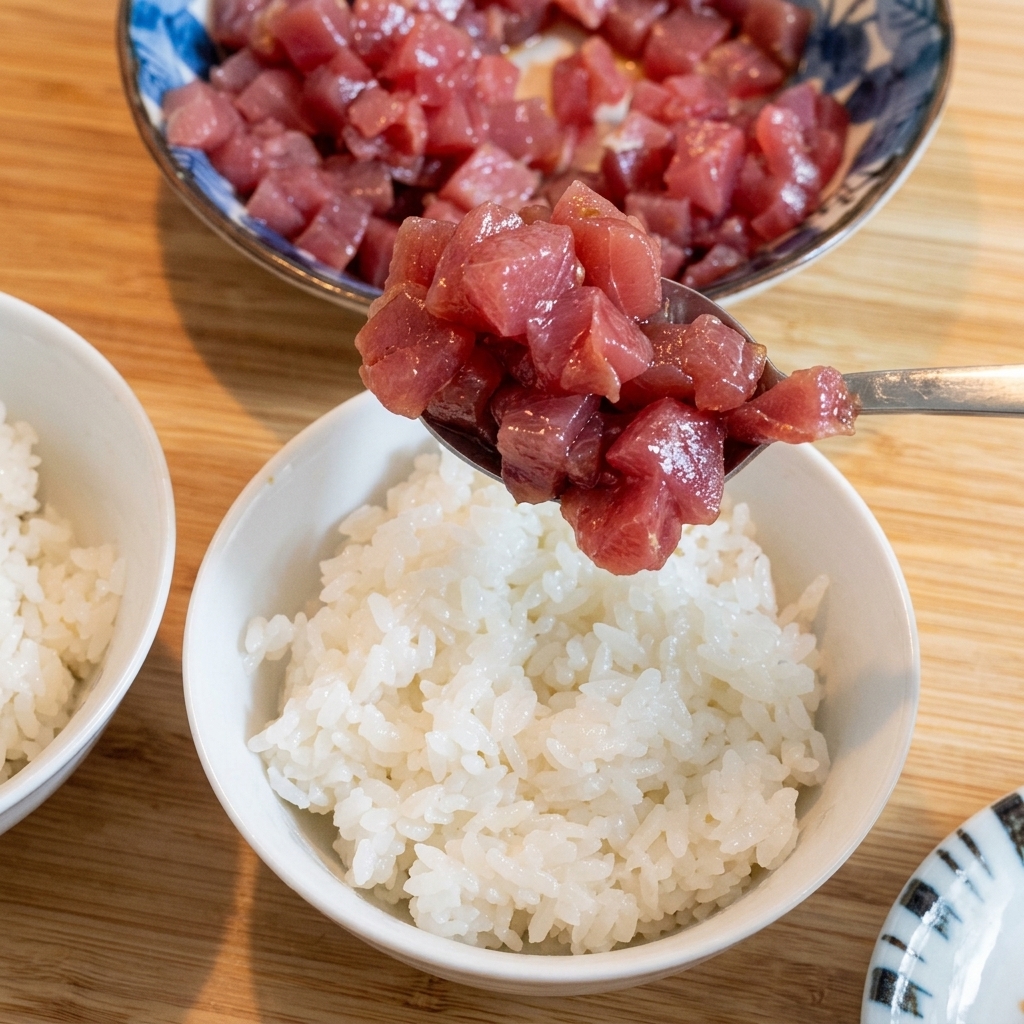 Spoonful of marinated tuna being carefully layered on top of white rice.