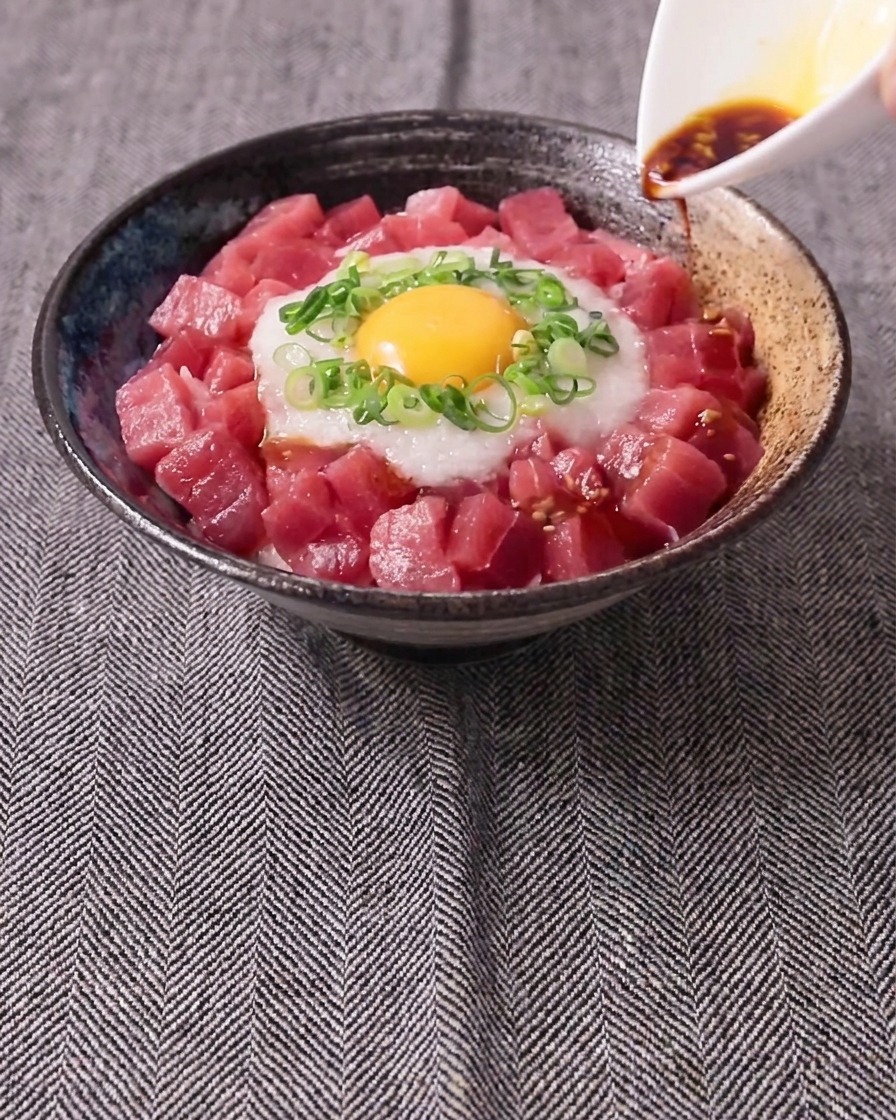 A dark sauce being poured from a small white pitcher over a bowl of cubed raw tuna, white grated yam, scallions, and a raw egg yolk.