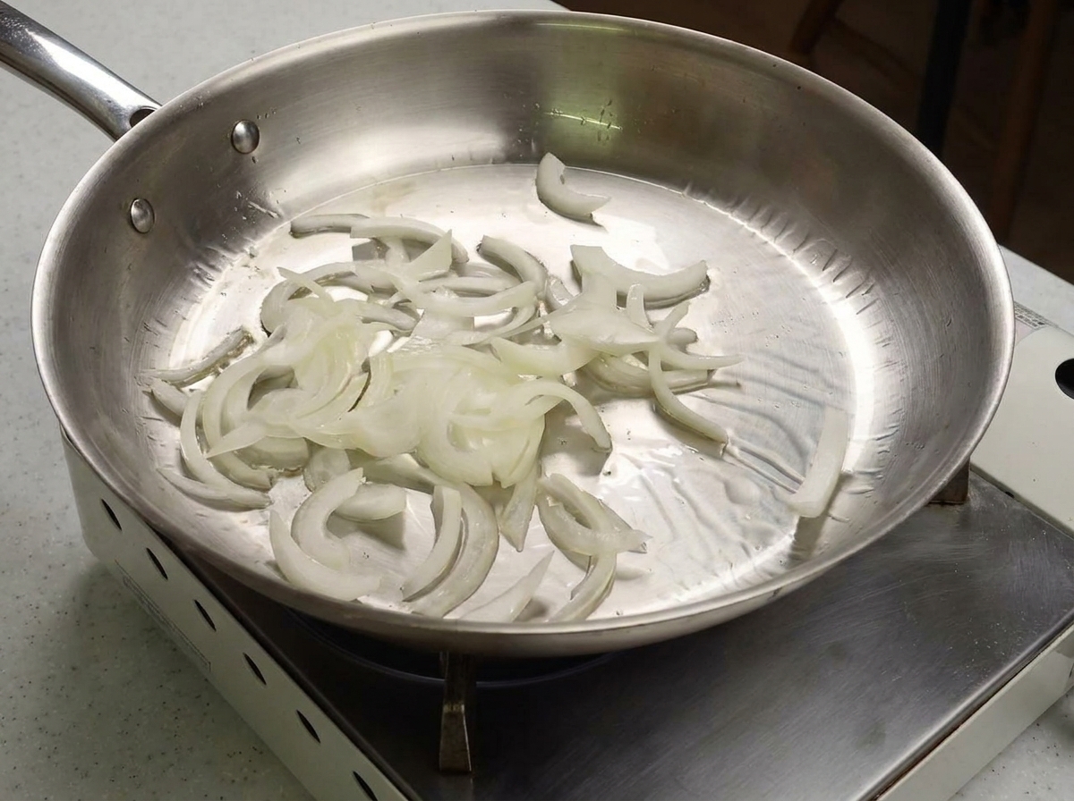 Slices of white onion being sautéed in a shiny stainless steel pan.
