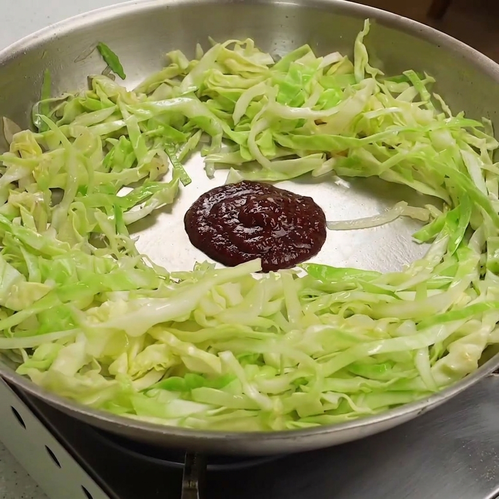 A dark red dollop of mixed chili and soybean paste placed over softened shredded cabbage in a pan.