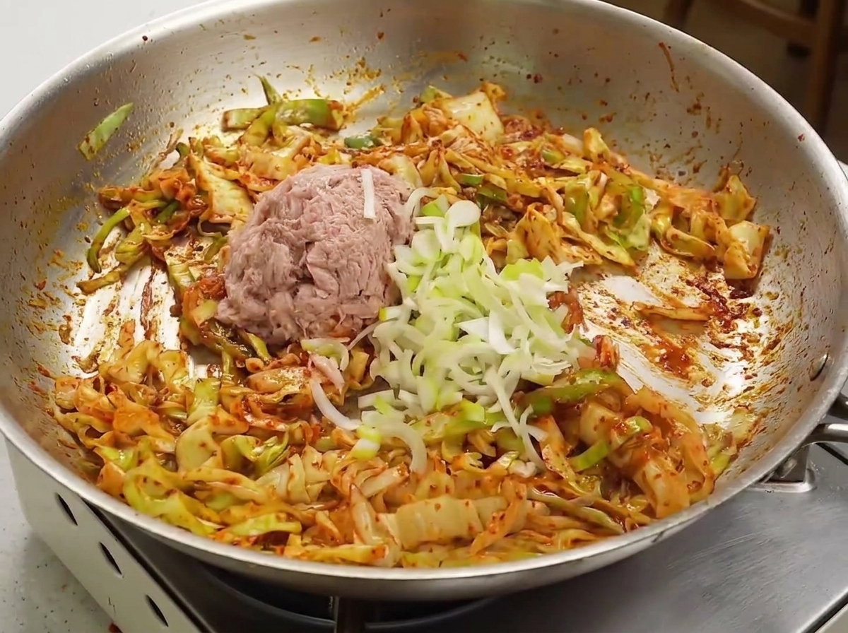 Flaked canned tuna and chopped green onions resting on top of a red, sauce-coated cabbage mixture in a pan.