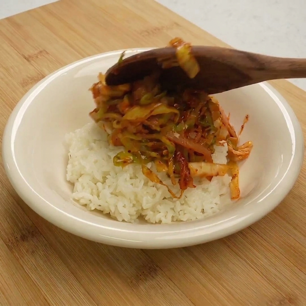 A wooden spoon serving the finished tuna and cabbage stir-fry over a mound of white rice in a white bowl.