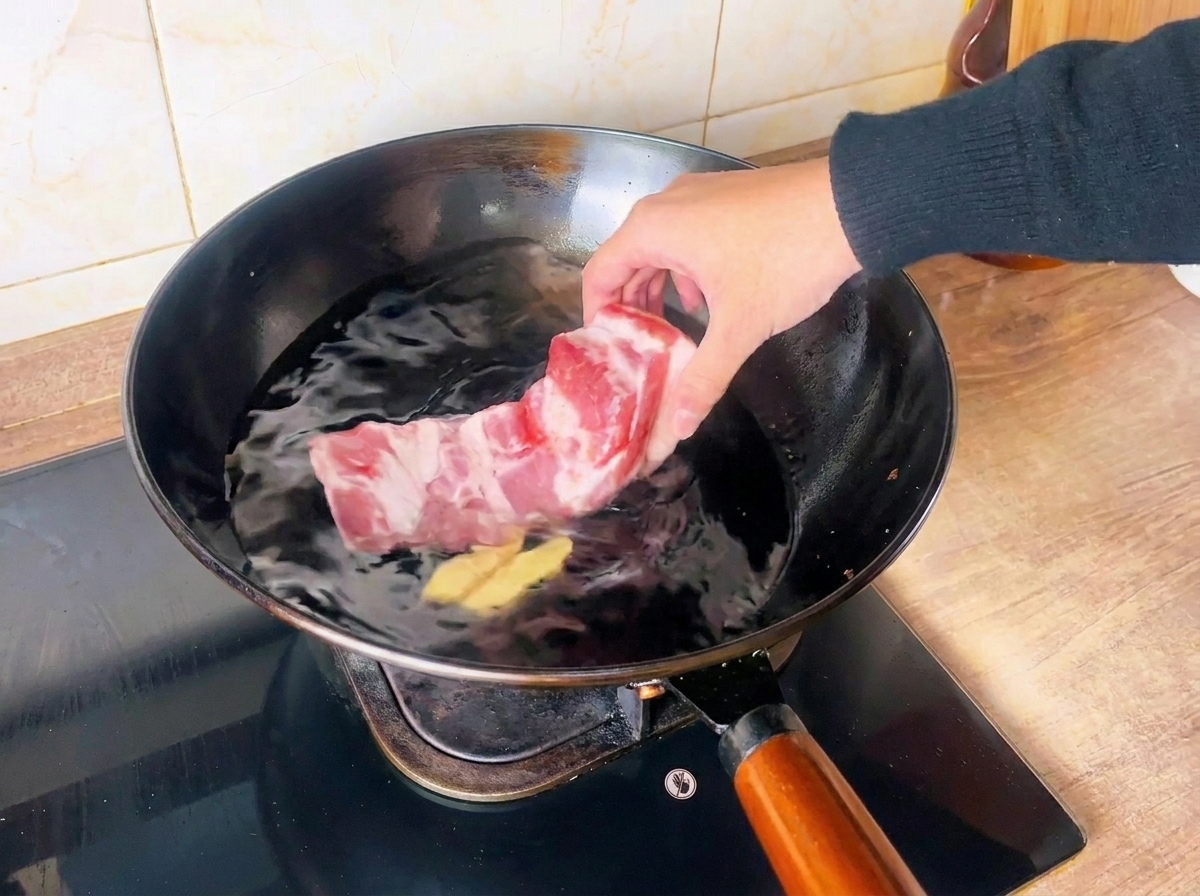 A hand placing a raw pork belly block into a wok filled with cold water and ginger slices.