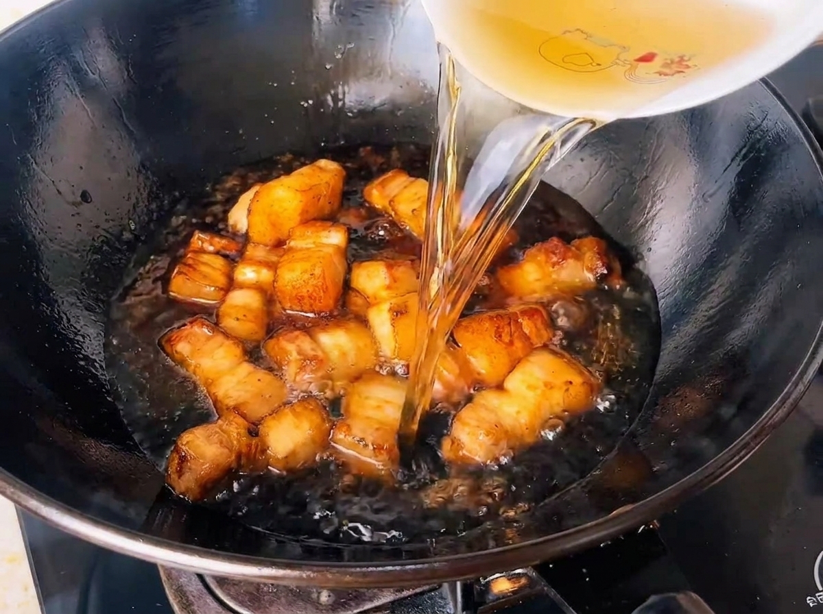 Steeped green tea water being poured into a wok full of seasoned pork belly cubes.