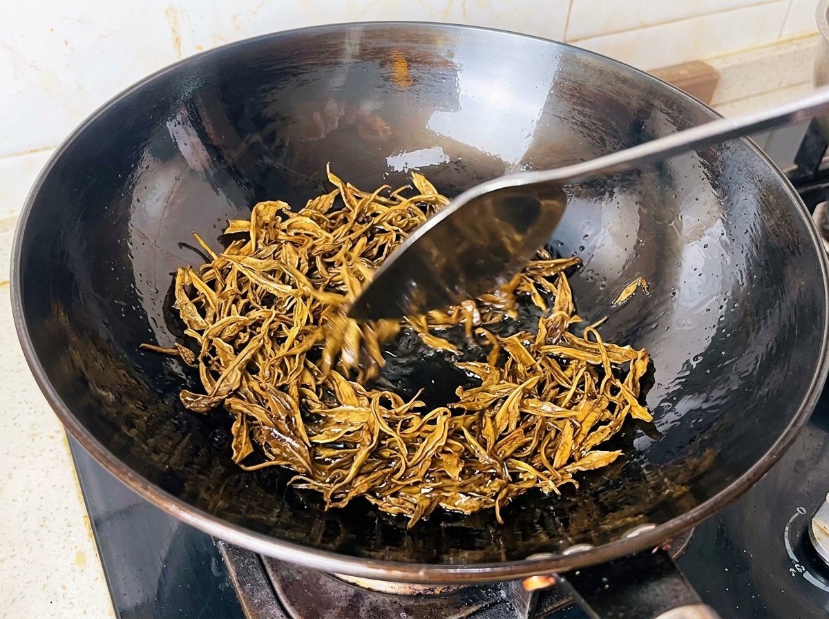 Wet green tea leaves being fried in hot oil inside a black wok with a metal spatula.