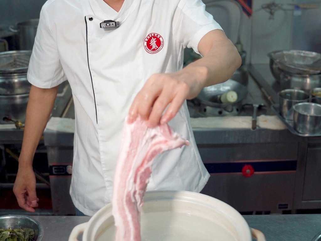 A chef holding a large piece of raw pork belly over a white pot filled with water.