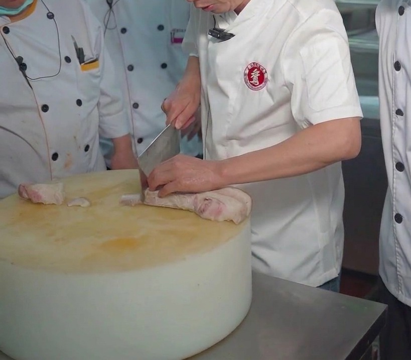 A chef using a cleaver to slice a block of boiled pork belly on a large, thick round cutting board.