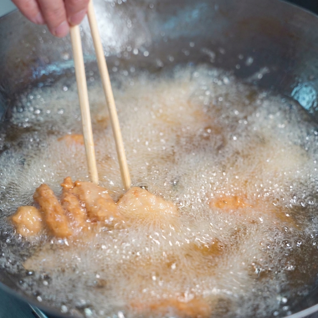 Coated pork pieces frying in bubbling golden oil, being stirred with wooden chopsticks.