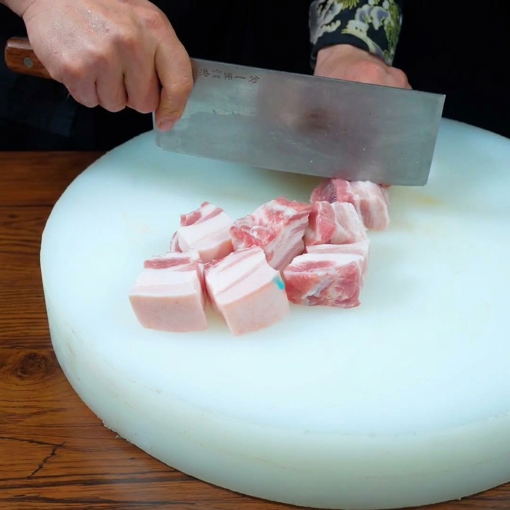 Hands using a cleaver to cut a slab of three-layer pork belly into uniform cubes on a white cutting board.