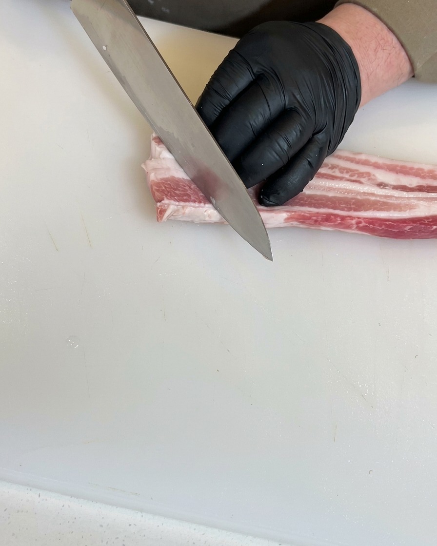 Hands wearing black gloves using a chefs knife to make deep diagonal cuts into a piece of raw pork belly on a white cutting board.
