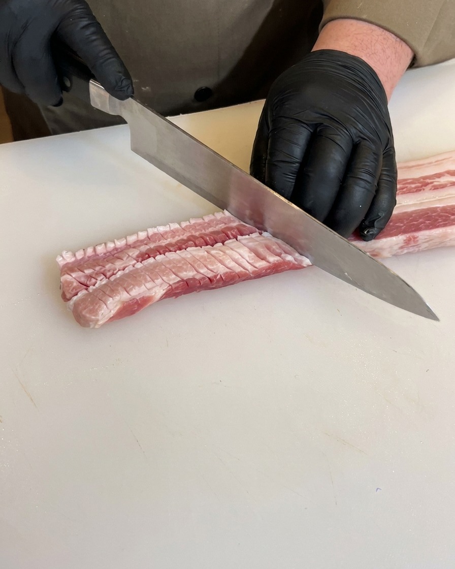 Hands in black gloves scoring the reverse side of a raw pork belly with a chefs knife, showing deep grid-like cuts along the meat.