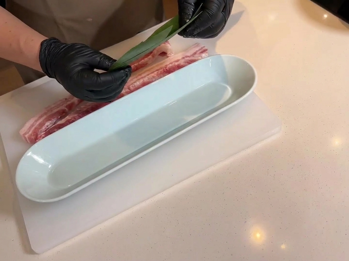 Hands holding a green bamboo leaf over a long white serving tray, with the scored raw pork belly resting nearby on the cutting board.