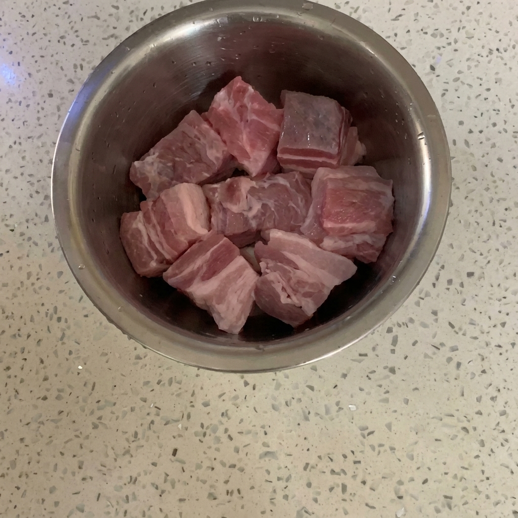 Raw pork belly cubes sitting in a stainless steel mixing bowl on a speckled countertop.