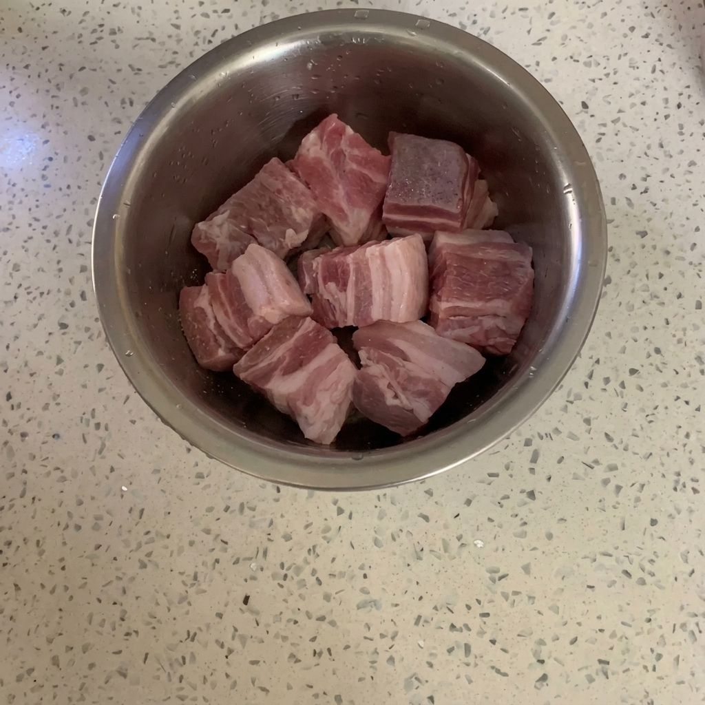 A metal bowl containing cubed raw pork belly being sprinkled with pepper powder.
