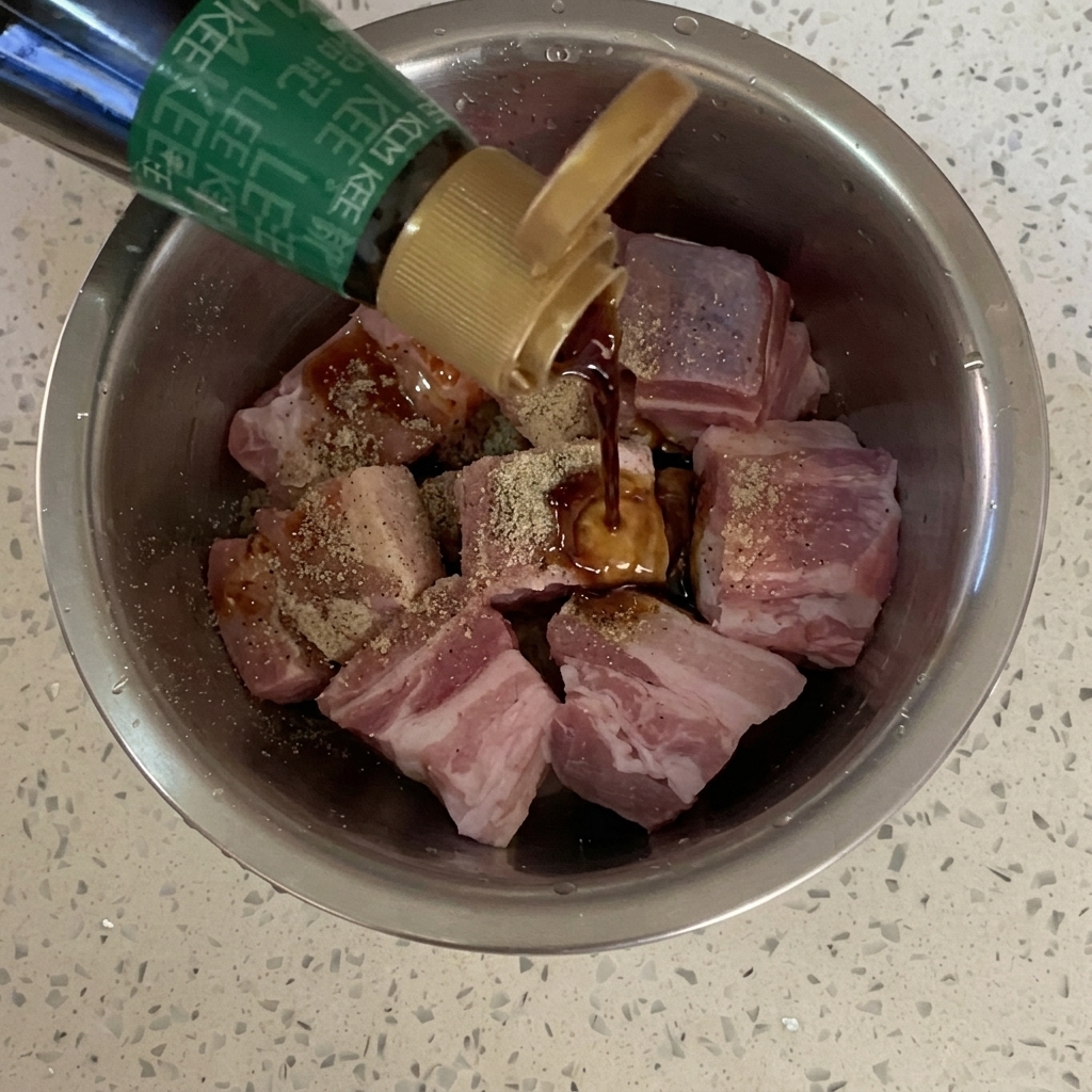 Soy sauce being poured from a bottle onto seasoned pork belly cubes in a metal bowl.