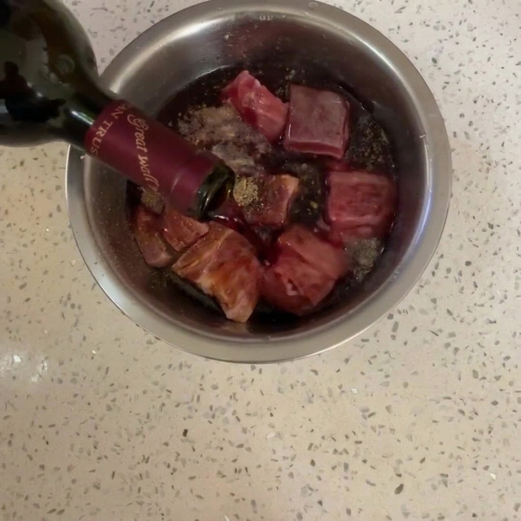 Red wine being poured from a dark glass bottle over seasoned pork belly in a stainless steel bowl.