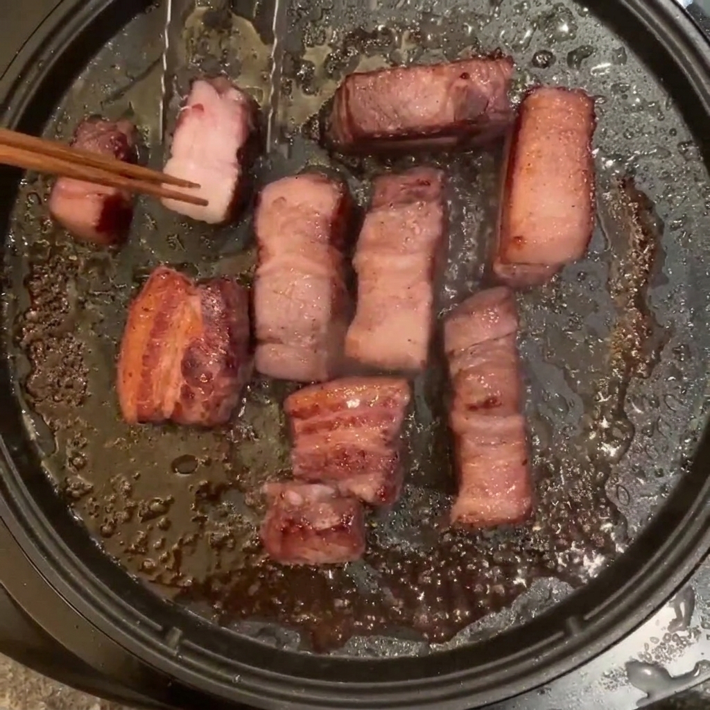 Several pieces of pork belly frying in an electric pan, being turned with wooden chopsticks.