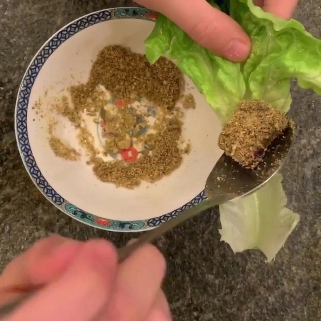 A close-up of a spoon holding a piece of cooked pork belly coated in crushed peanuts, hovering over a romaine lettuce leaf.