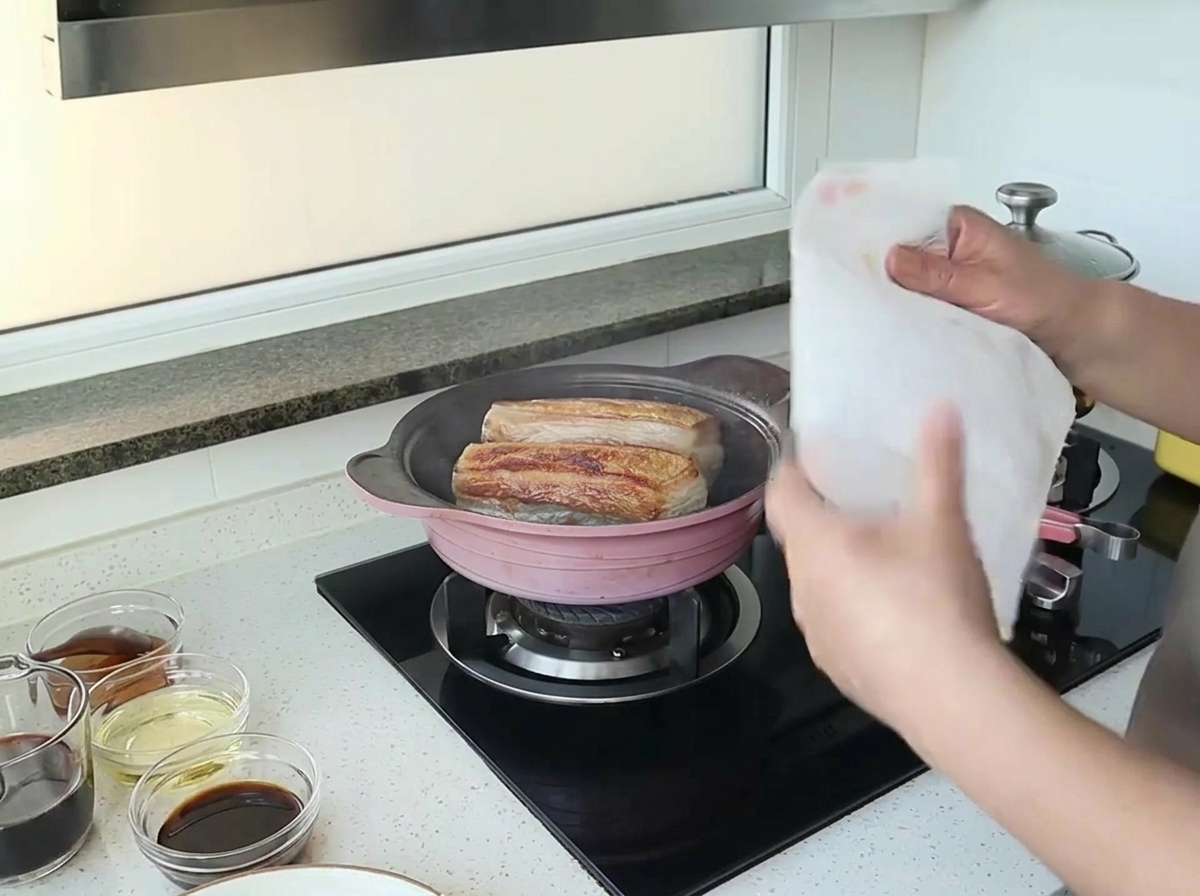 Hands holding a white kitchen paper towel over a pan with two pieces of searing pork belly.