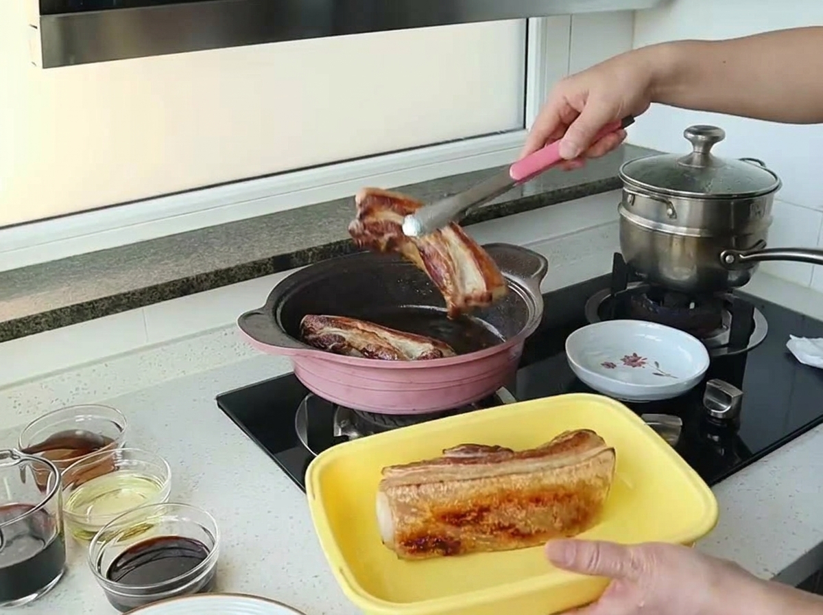 Tongs lifting a beautifully browned piece of pork belly out of a pink cooking pan.