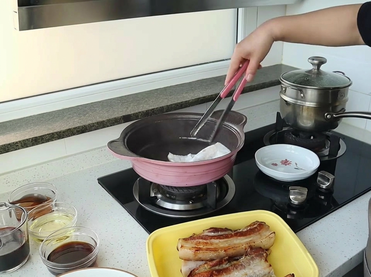 A hand using tongs to hold a folded paper towel, wiping excess oil from the inside of a pink pan.