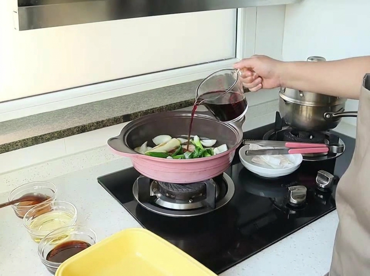 A hand pouring red wine from a glass measuring cup into a pan filled with sautéed onions and aromatics.