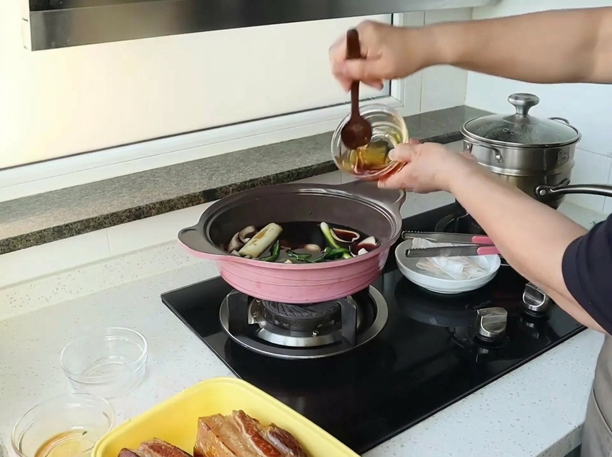 A hand pouring honey from a small glass bowl into a pink pot filled with a dark liquid mixture and aromatics.