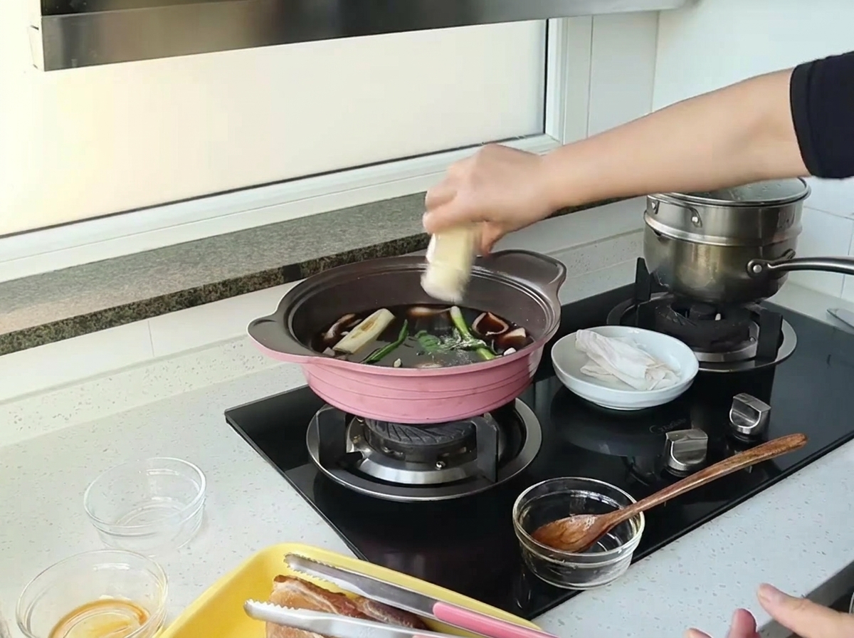 A hand shaking ginger powder from a small jar into a pot with dark sauce, onions, and green onions.