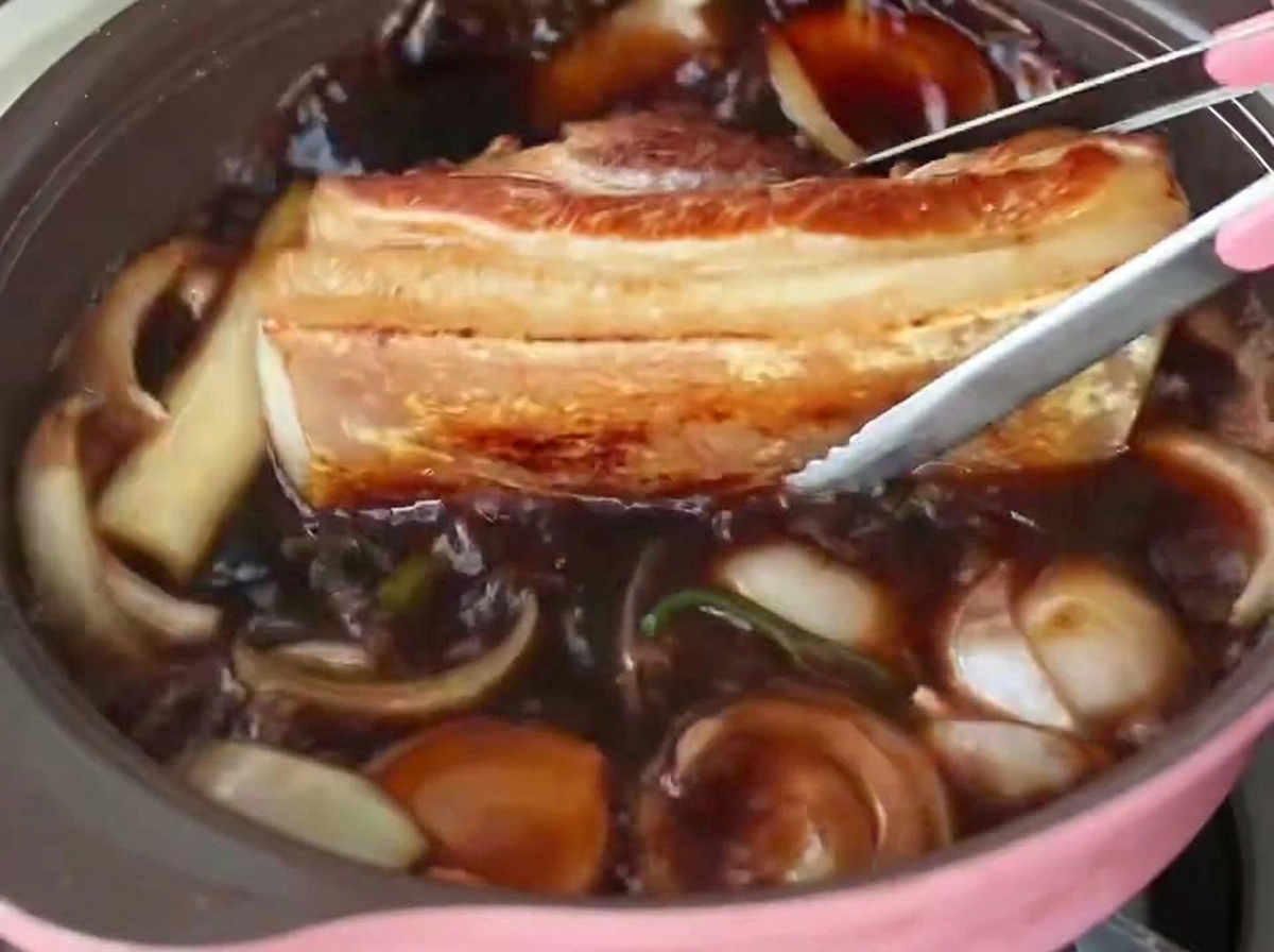 Tongs placing a thick strip of seared pork belly into a pot of dark, simmering braising liquid with onions.
