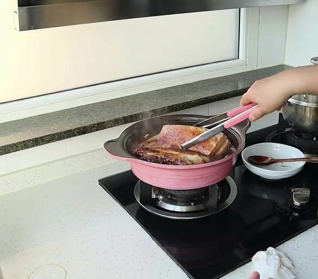 A person using tongs to flip large pieces of pork belly simmering in a dark sauce in a pink pot.