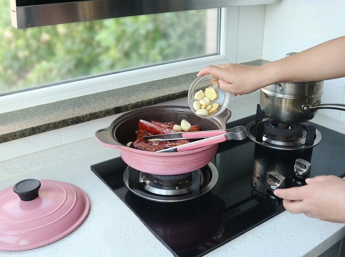 A hand pouring peeled garlic cloves and a slice of ginger from a small glass bowl into a pink pot containing dark, braised pork belly.