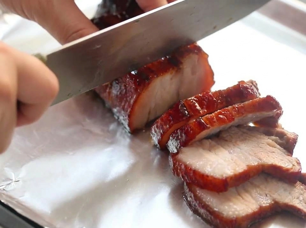 A chefs knife slicing through a piece of dark, glazed red wine pork belly resting on a piece of aluminum foil.