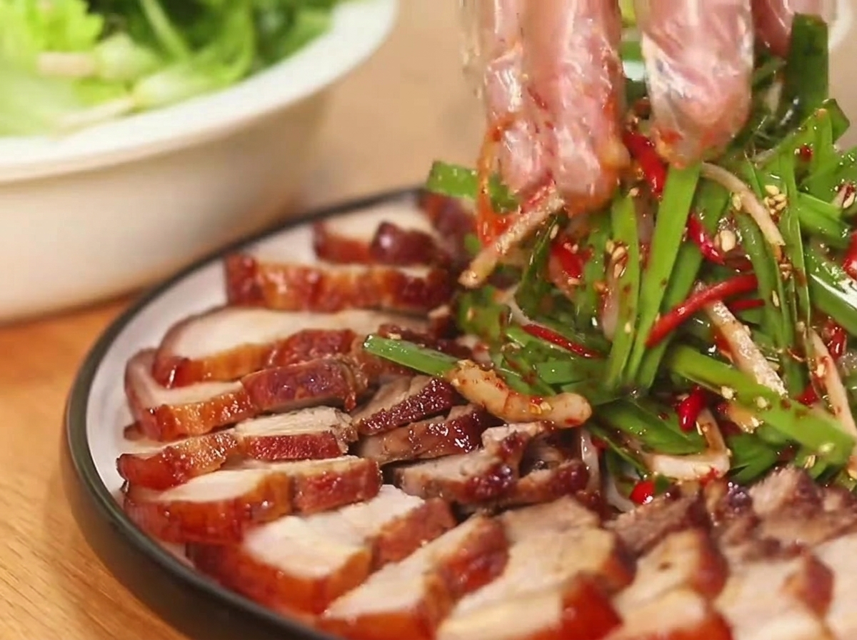 Gloved hands tossing a vibrant green scallion and red chili salad next to sliced glazed pork belly arranged on a round plate.
