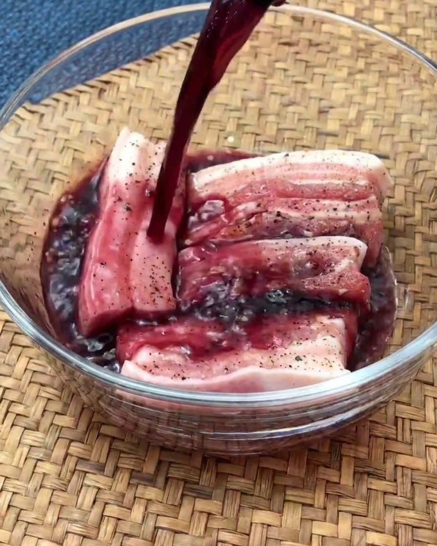 Raw pork belly slices marinating in a glass bowl filled with red wine.