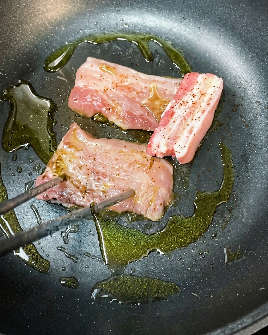 Marinated pork belly slices browning in a hot pan with olive oil.