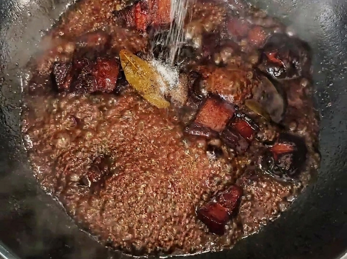 Salt being sprinkled over simmering pork belly cubes in a dark, rich sauce.