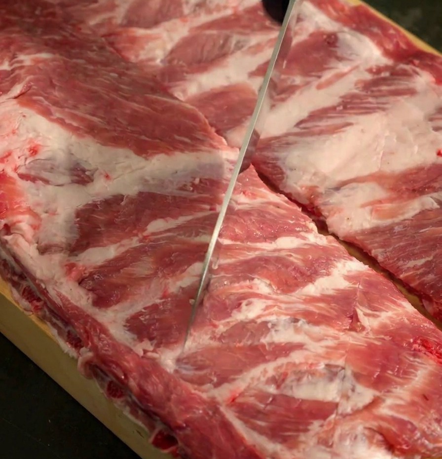 A sharp knife scoring the surface of two raw racks of pork ribs on a wooden cutting board.