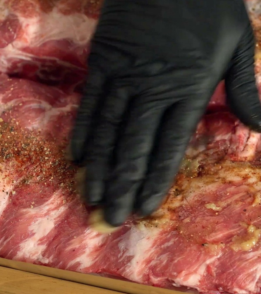 A hand in a black glove rubbing spices and garlic into raw pork ribs.