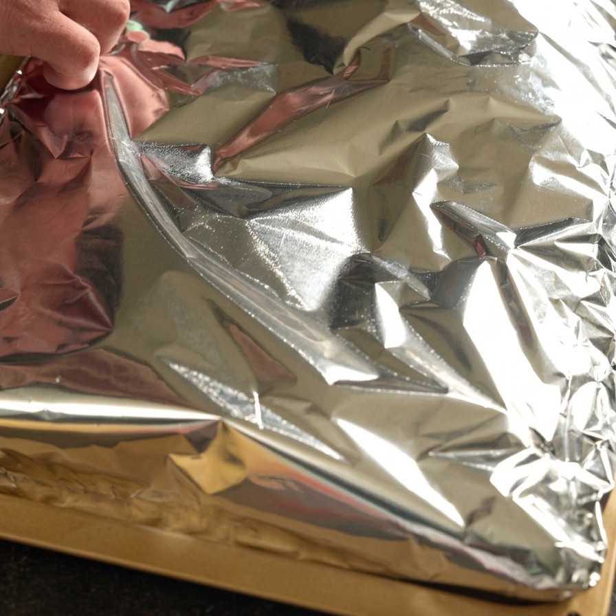 Hands folding aluminum foil tightly over a rack of seasoned pork ribs.