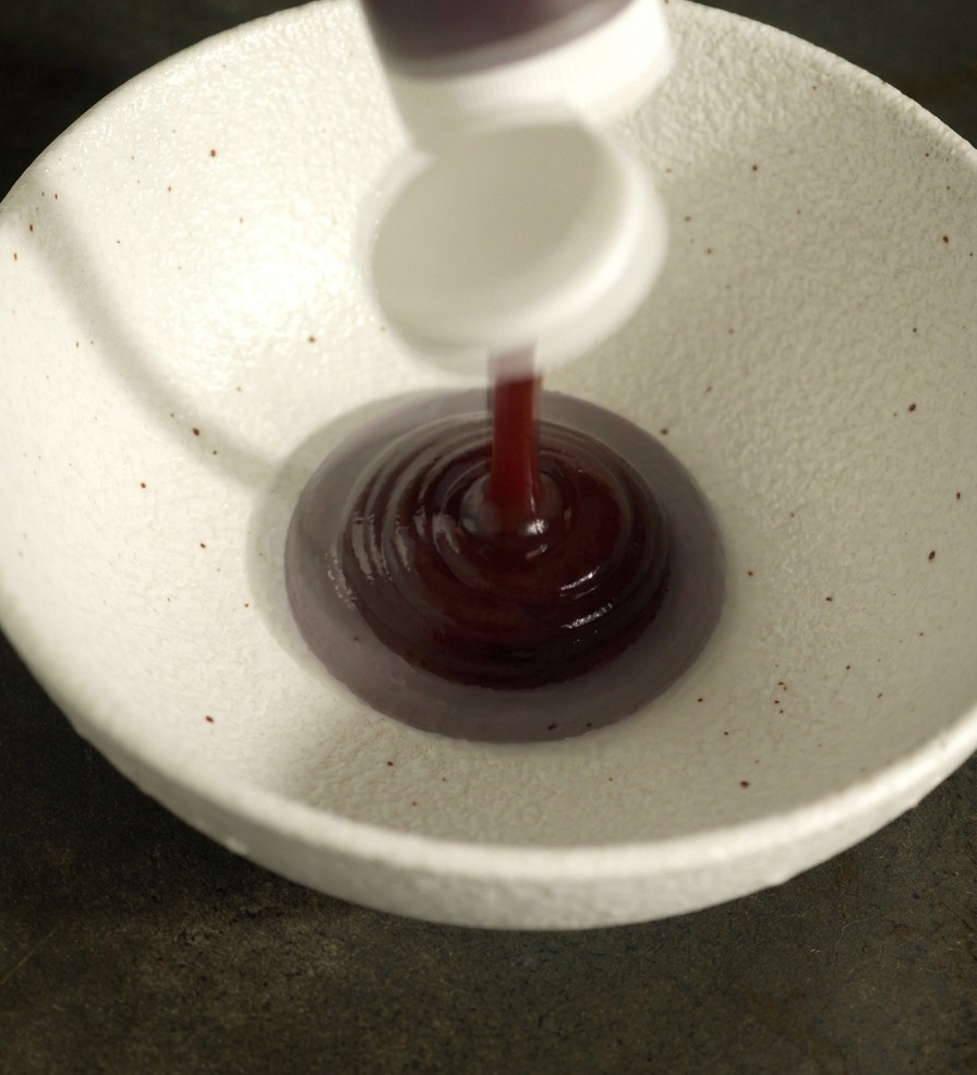 Dark red barbecue sauce and other liquid seasonings being poured into a textured white ceramic bowl to create a glaze.