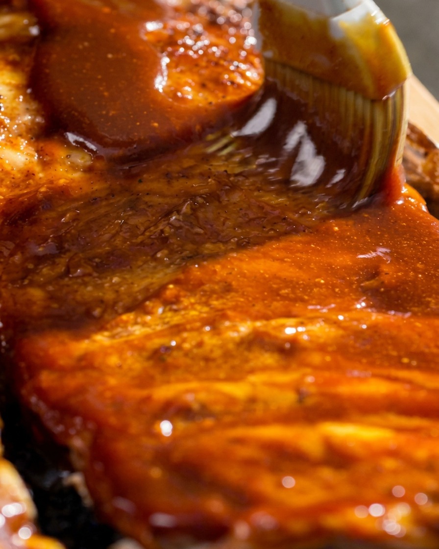A close-up shot of a pastry brush spreading thick, glossy red barbecue glaze across the surface of cooked ribs.