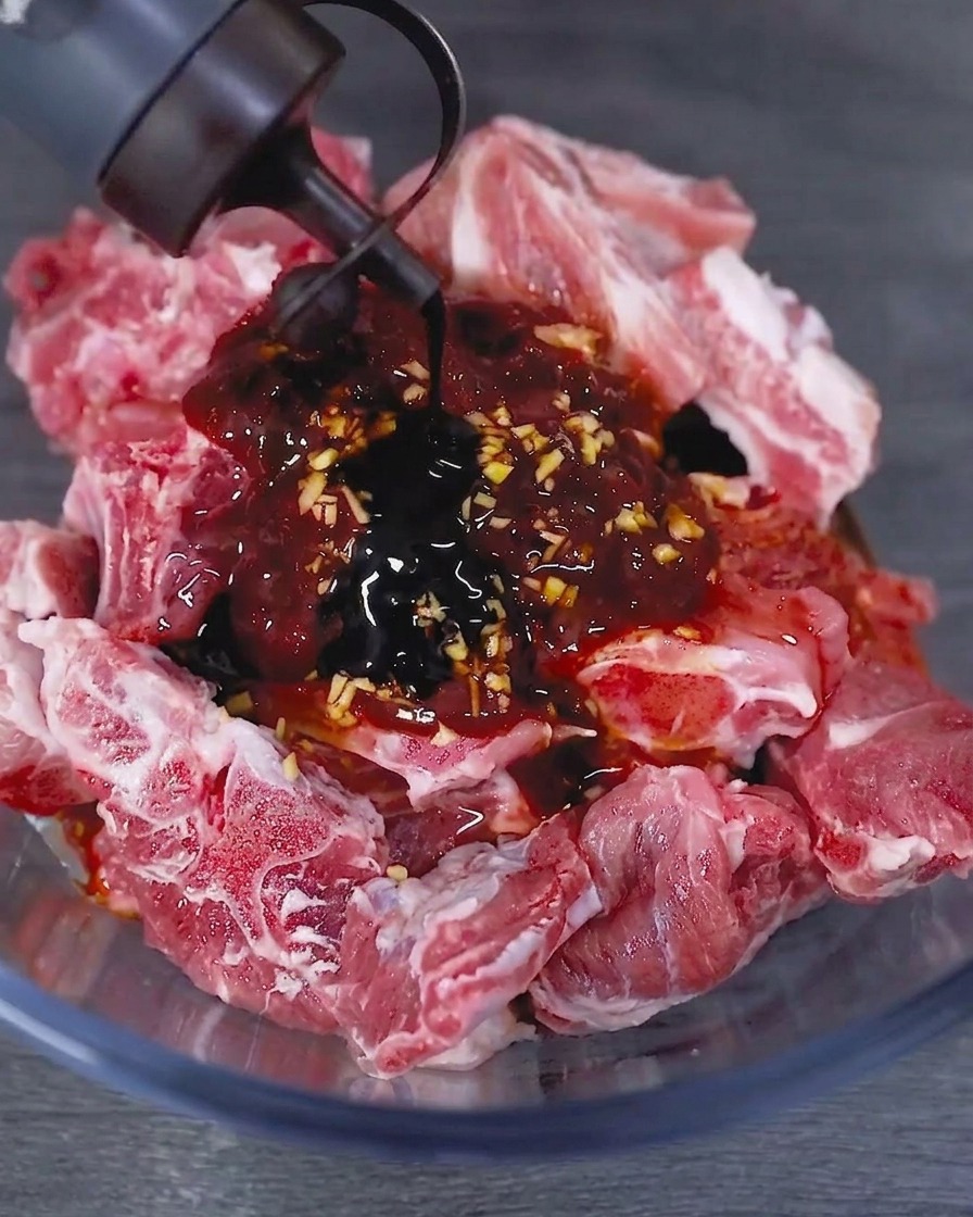 Dark soy sauce being poured from a bottle into a glass bowl containing marinated raw pork ribs.