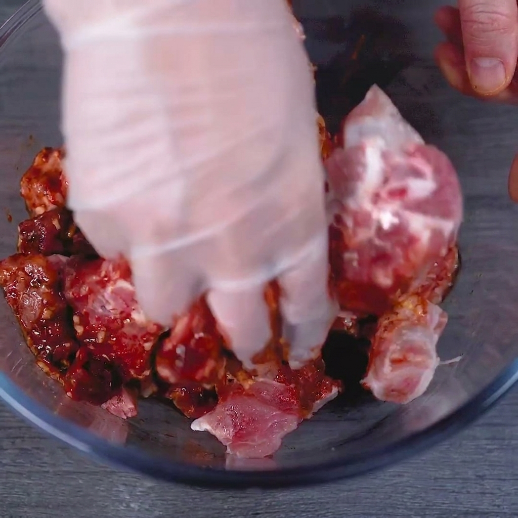 A gloved hand thoroughly massaging the red marinade into the raw pork ribs in a glass bowl.
