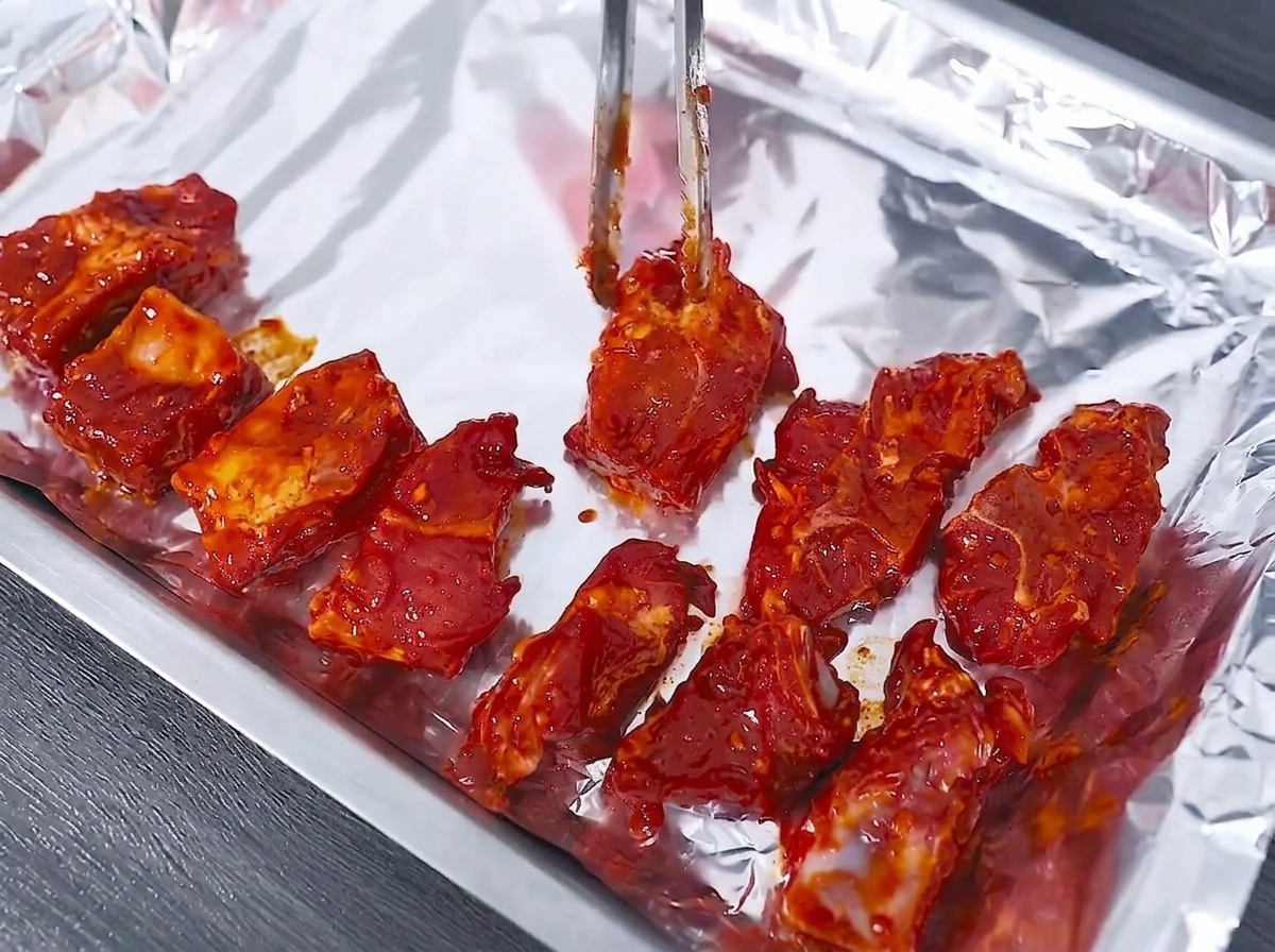 Raw marinated pork ribs being neatly arranged on an aluminum foil-lined baking tray using metal tongs.