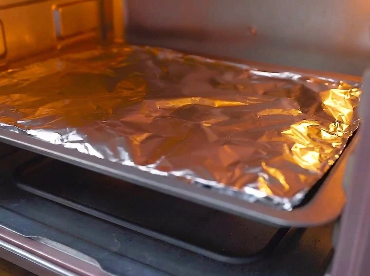 A foil-covered baking tray being placed on the middle rack of a preheated, glowing oven.