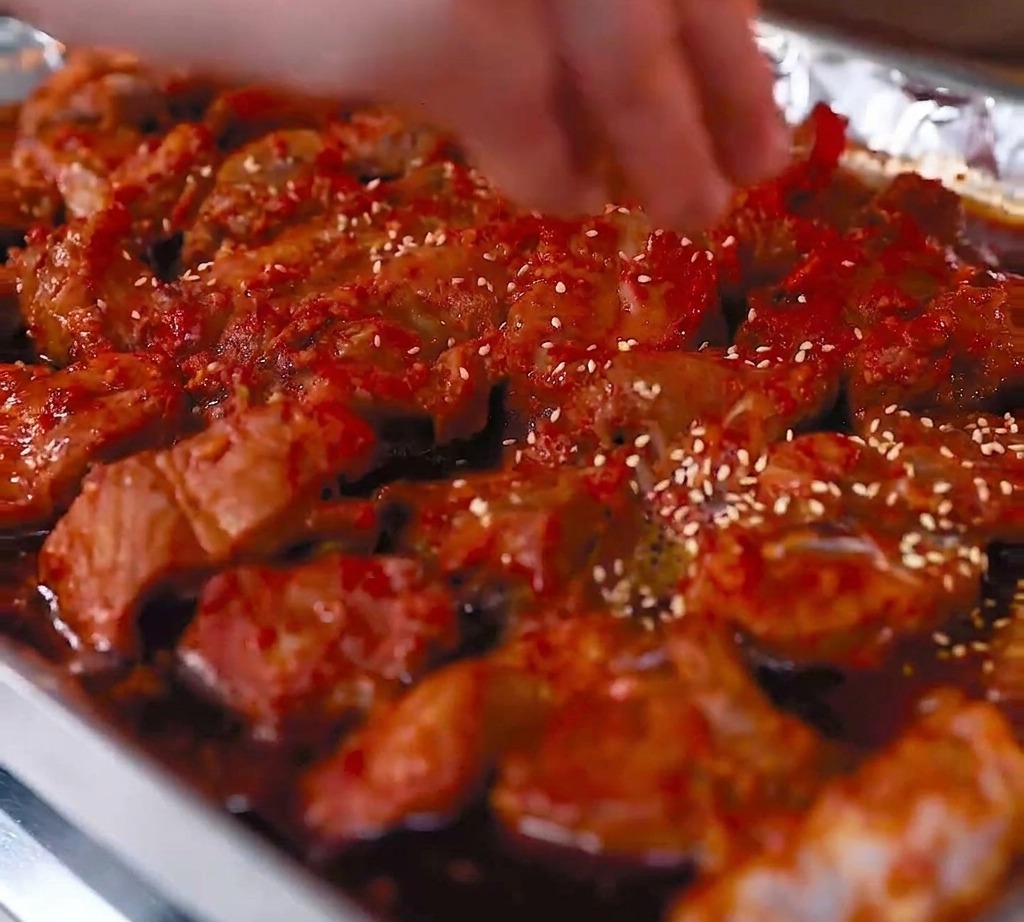 A close-up view of roasted pork ribs in a foil-lined tray, with white sesame seeds being sprinkled over the glistening, red-glazed surface.