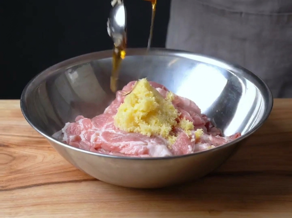 Raw sliced pork in a metal bowl being combined with lemon zest and corn syrup.