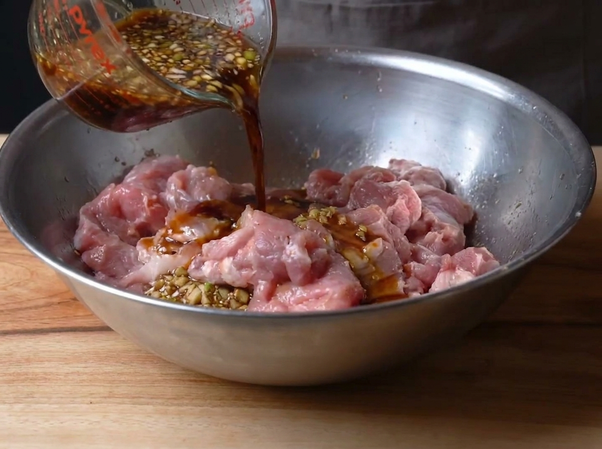 The dark, savory marinade being poured over the pre-marinated pork in a metal bowl.